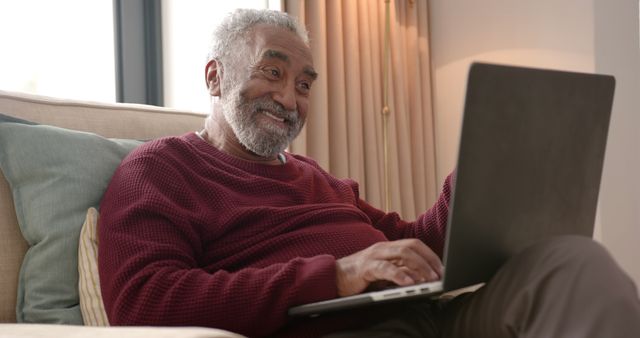 Senior man sitting on couch using laptop, smiling and enjoying leisure at home. Ideal for advertisements and promotions related to senior living, technology for elderly, home comfort, and retirement lifestyle.