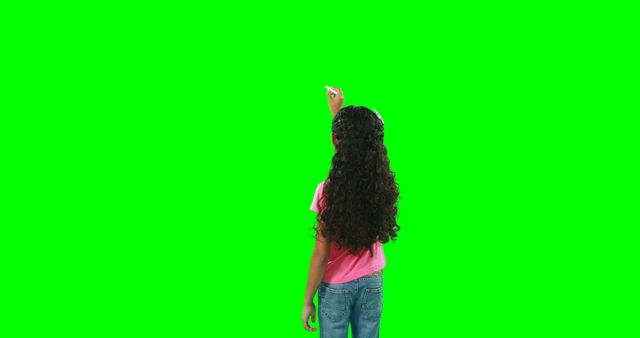 A young girl with long curly hair is standing in front of a green screen, pointing upwards, with copy space. Her casual attire and the green screen background suggest a setting that's ready for digital editing or visual effects.