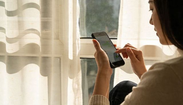 Asian woman tapping smartphone by sheer curtain window in warm sunlight, texting