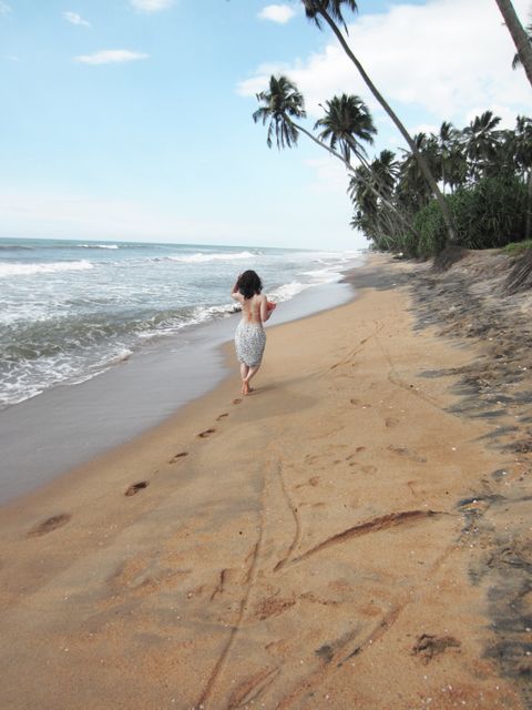 Woman walking along a serene tropical beach with palm trees and ocean waves in the background. Footprints are visible in the sand, creating a peaceful vacation vibe. Perfect for travel advertisements, beach holiday brochures, or relaxation themed articles. Ideal for promoting tropical destinations or beach activities.