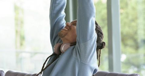 Mid-adult man stretching on sofa with pink headphones by floor-to-ceiling windows, relaxed morning