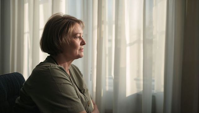 Senior woman gazing through sheer curtains in sunlit room, contemplative home portrait