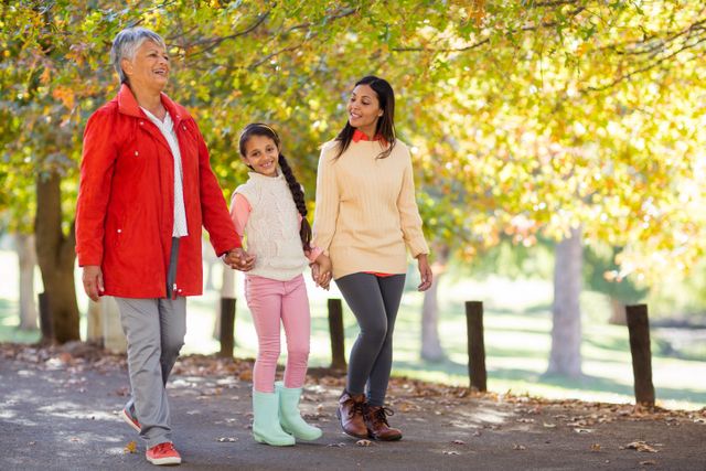 Three generations of women, including a grandmother, mother, and daughter, walking together in a park during autumn. They are holding hands and smiling, enjoying a family outing. This image can be used for promoting family values, outdoor activities, and multigenerational bonding. Ideal for advertisements, family-oriented content, and health and wellness campaigns.