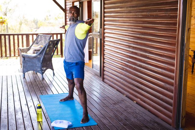 Bald african american senior man with arms outstretched exercising on mat in log cabin. Confident, unaltered, vacation, retirement, solitude, yoga, beard, fitness and active lifestyle concept