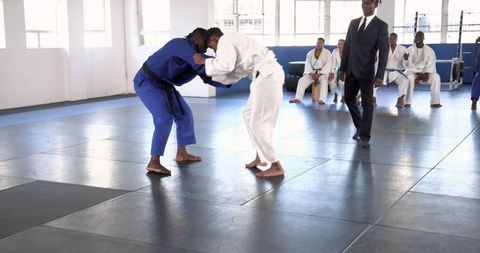 Martial arts competitors grappling in gym with referee overseeing match