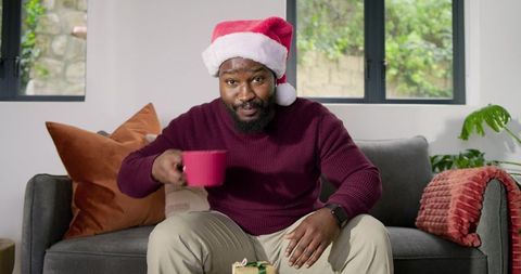 African american man wearing santa hat holding red mug sitting on cozy sofa celebrating holidays