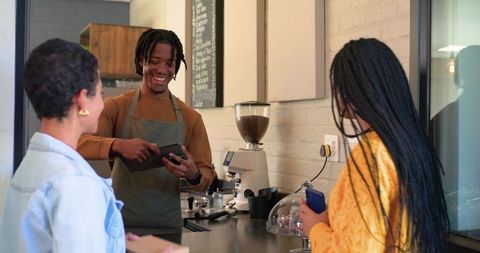 Smiling barista serving customers using tablet while taking orders and mobile payments