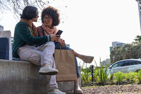 Friends sharing smartphone during shopping break in urban park, casual lifestyle moment