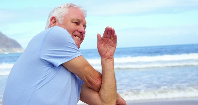 An elderly man wearing a light blue shirt is stretching his arms while standing close to the shoreline. The ocean waves and a hint of the coastline are visible in the background. Suitable for wellness content, fitness advice for seniors, healthy aging campaigns, and active lifestyle promotions.
