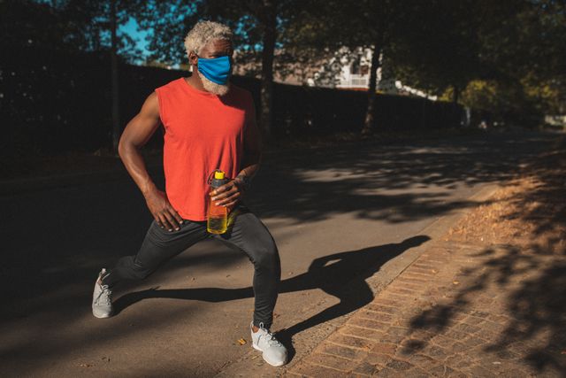 African american senior male exercising outdoors wearing mask stretching in sunny street. healthy active retirement lifestyle during coronavirus covid 19 pandemic.