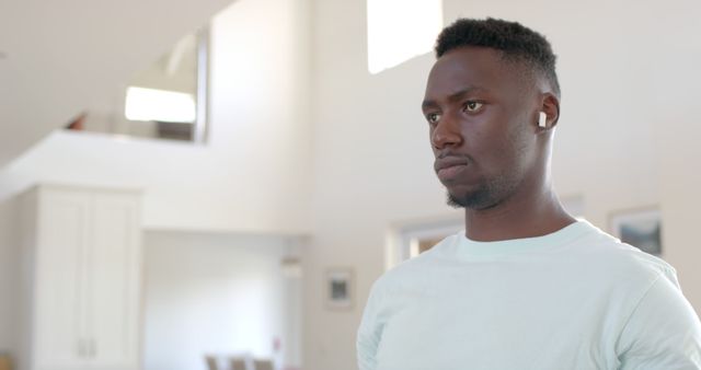 The young man is concentrating while wearing wireless earbuds in a modern living space. This can be used for advertisements or articles on technology, modern lifestyles, or focused activities at home.