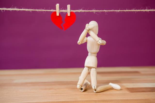 This image depicts a wooden figurine kneeling in front of a broken heart hanging on a string with clothespins. The scene conveys themes of heartbreak, sadness, and emotional distress. Ideal for use in articles, blogs, or social media posts about relationships, breakups, and emotional well-being.