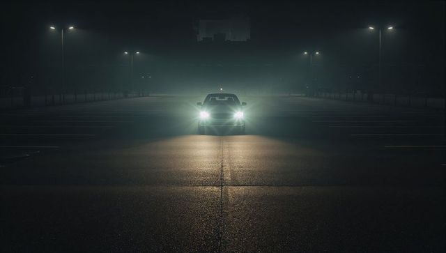 Lonely car with bright headlights piercing fog in empty wet parking lot at night