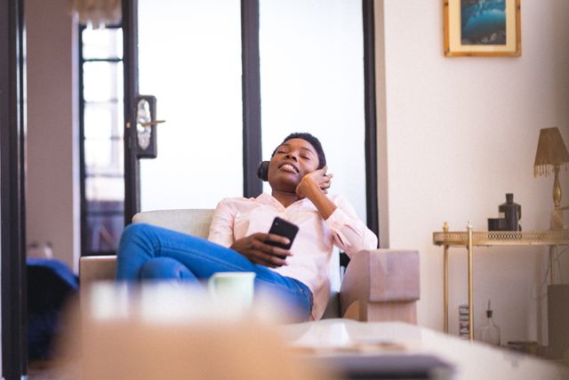 Young African American woman sitting comfortably at home, holding a smartphone and listening to music through headphones. Ideal for use in lifestyle blogs, technology advertisements, or articles about modern living and relaxation.