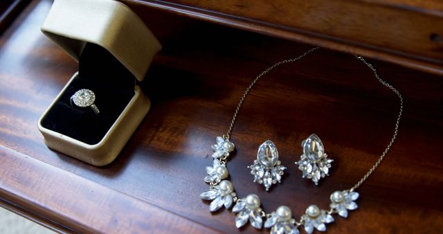 Elegant arrangement of a diamond ring in a jewelry box, a floral pearl and diamond necklace, and matching earrings on a varnished wooden surface. Suitable for themes of luxury, fashion, wedding accessories, engagement, gift pieces, vintage collections, and feminine elegance.