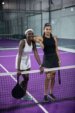 Young black and south asian women posing at net on vibrant purple padel court wearing skirts