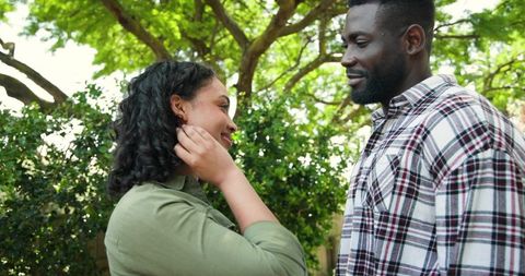 Smiling couple touching hair and gazing under sunlit tree, intimate backyard romance