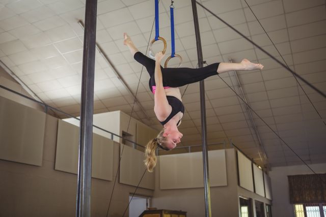 Female gymnast practicing on rings in a gymnasium, showcasing strength, balance, and flexibility. Ideal for use in articles or advertisements related to gymnastics, athletic training, fitness, sports, and exercise. Can also be used for motivational content highlighting determination and dedication in sports.