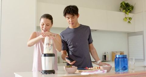 Couple making smoothie at kitchen island, woman securing blender jar, man checking smartphone
