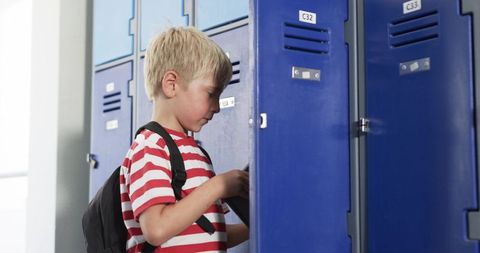 Blond schoolboy opening locker in school corridor wearing striped shirt and backpack