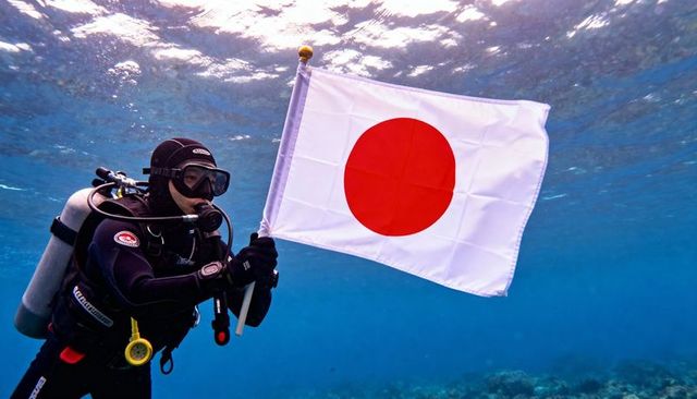 Scuba diver holding japan flag underwater near coral reef, sunlit ocean exploration