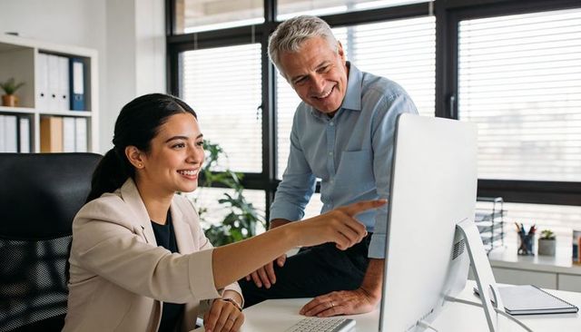 Young professional woman pointing at desktop monitor while colleague reviewing project