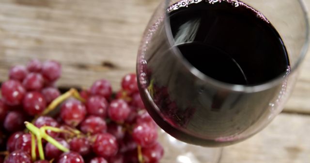 A glass of red wine is tilted on a wooden surface near a bunch of red grapes, with copy space. The scene suggests a tasting or pairing of wine with fresh grapes, evoking a sense of indulgence and the pleasures of viticulture.