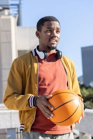 African american man holding basketball wearing headphones on sunlit rooftop