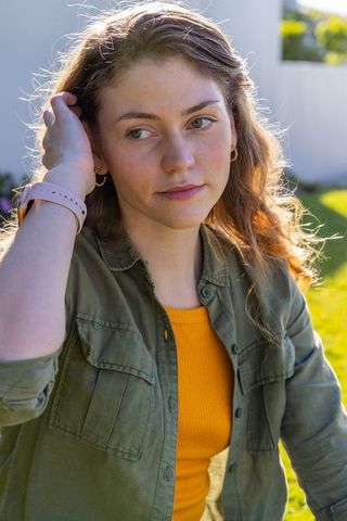 Contemplative young woman sitting in garden touching hair wearing mustard top olive jacket