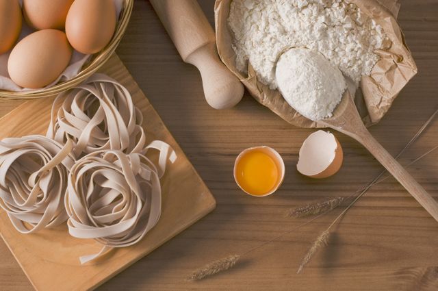 This image depicts fresh homemade pasta ingredients on a rustic wooden table. It includes eggs, a bag of flour with a wooden spoon, a rolling pin, and neatly arranged pasta nests. Ideal for food blogs, cooking tutorials, recipe books, or culinary articles about fresh homemade cooking or Italian cuisine.
