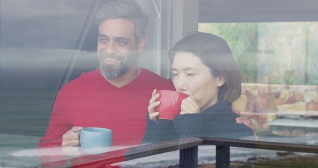 Interracial couple is enjoying a peaceful morning together, drinking coffee near a large window. The setting exudes a cozy, relaxed atmosphere, perfect for illustrating concepts of togetherness, bonding, and enjoying simple pleasures. This image is suitable for use in lifestyle blogs, health and wellness advertisements, and social media posts focusing on relationships and mindfulness.