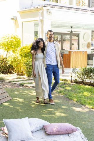Young couple walking arm-in-arm through sunlit backyard near blanket with pillows