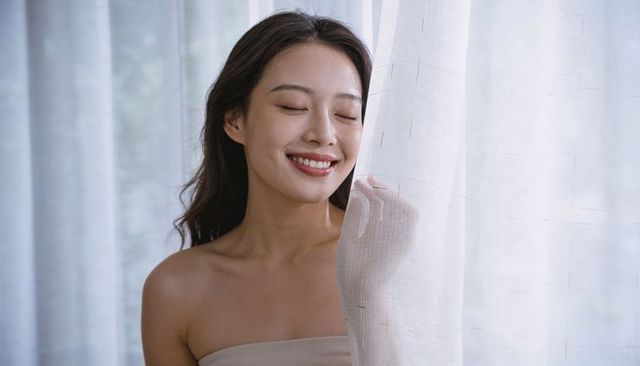 Asian woman smiling with eyes closed touching sheer curtain in serene minimalist interior