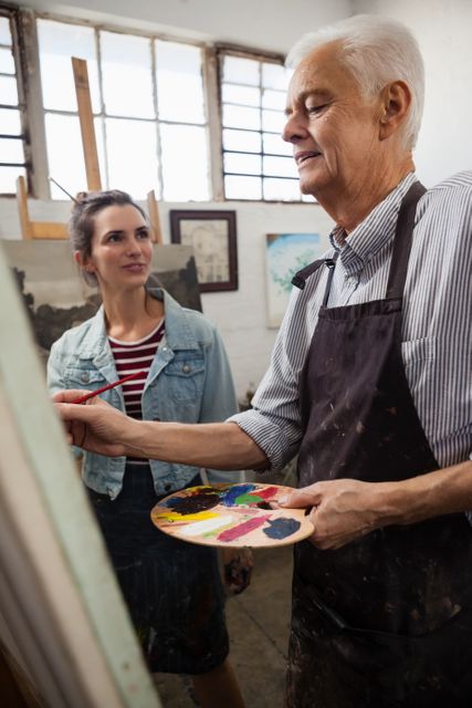 Senior man painting on canvas while woman interacts in art class. Ideal for themes of creativity, learning, mentorship, and artistic expression. Suitable for use in educational materials, art class promotions, and content related to hobbies and senior activities.