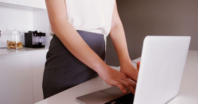 Businesswoman standing while working on laptop at home kitchen counter. Ideal for concepts related to remote work, productivity, home office setups, work-life balance, and modern workplaces.