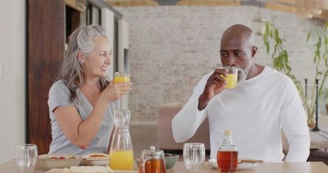 Senior couple enjoying sunlit breakfast at home sipping orange juice with pancakes