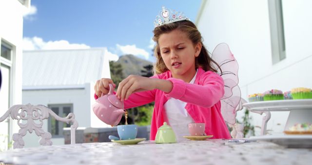 A young Caucasian girl dressed as a fairy princess is pouring tea at a playful outdoor tea party, with copy space. Her expression suggests she is deeply engaged in imaginative play, enhancing the whimsical atmosphere of the scene.