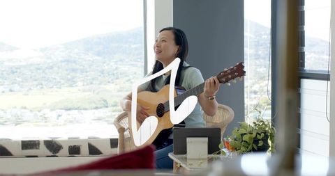 Asian woman playing acoustic guitar in sunlit lounge by large window with tablet