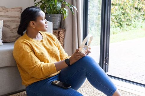 African american woman sitting by patio door holding headphones and smartphone