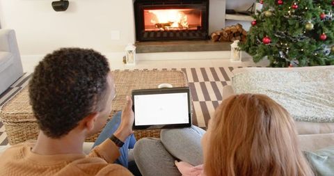 Diverse couple sitting on couch holding tablet by fireplace with christmas tree