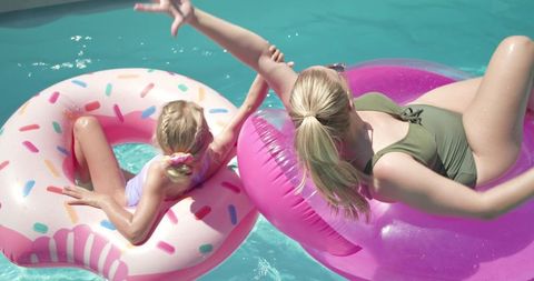 Mother and daughter floating on pink donut and tube in sunlit turquoise pool