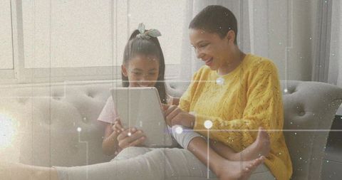 Mother and daughter sharing tablet, laughing and pointing at screen on sunlit cozy sofa