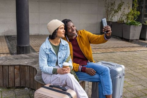 Multicultural couple taking selfie while waiting with carry-on luggage in urban plaza