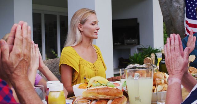 Family members sitting together around a table outdoors, holding hands, with eyes closed, praying before meal. Fresh fruits, lemonade, and bread rolls on the table. Ideal for concepts like family values, gratefulness, outdoor dining, and summer gatherings.