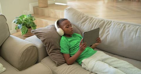 African-American Boy Relaxing on Sofa Wearing Headphones Using Tablet for Learning