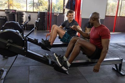 Male workout partners rowing on air rowers and fist bumping in bright gym