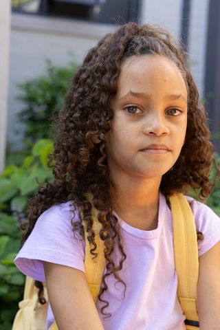 Young girl wearing yellow backpack and purple tee facing camera outdoors near city greenery
