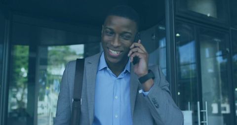 Smiling businessman talking on phone while exiting office glass doors with shoulder bag