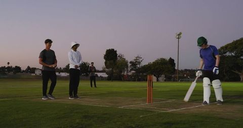 Batsman preparing at crease with teammates and umpire watching, dusk sky, lights glowing