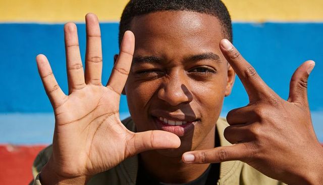 Winking male teen framing face with hands against colorful mural, playful streetstyle